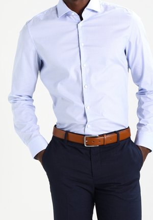 Mann trägt ein hellblaues Hemd mit Knöpfen, dunkelblaue Hosen und einen braunen Ledergürtel mit silberner Schnalle, die Hände in den Taschen.