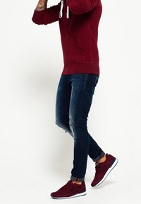 Sudadera con capucha color burdeos y cordones blancos, jeans de denim azul con rodilleras rasgadas y zapatillas burdeos con suelas blancas.