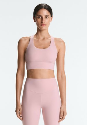 Femme aux cheveux foncés portant un soutien-gorge de sport rose clair et des leggings taille haute, debout face à un fond clair uni.