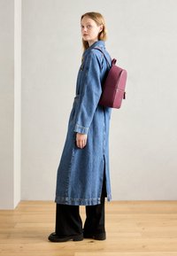 Denim lang frakk med belted midje og sidelommer, kombinert med en burgunder lærryggsekk med gullfarger og glidelåsdetailler.