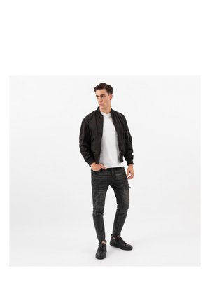 Uomo con giacca bomber nera, t-shirt bianca, jeans neri strappati e sneakers nere, in piedi con le mani nelle tasche su sfondo bianco.
