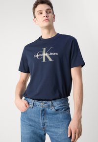 Camiseta de algodón en color navy con un gran logo plateado "CK Calvin Klein Jeans." Combinada con jeans de denim azul claro, mostrando un corte relajado y textura.