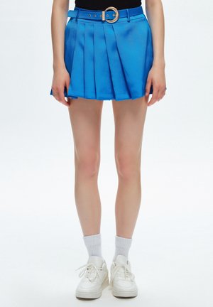 Blauwe geplooide rok met een gestructureerde glans, voorzien van een bijpassende riem met een ronde gesp, gestyled met witte sneakers en sokken tot halverwege de kuit.