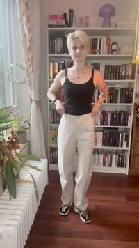 Une personne avec des cheveux blonds courts, portant un débardeur noir et un pantalon blanc, se tient dans une pièce avec des étagères, des plantes et un sol en bois.