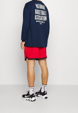 Personne portant une chemise bleu marine à manches longues avec le texte "National Basketball Association", un short rouge, des chaussettes blanches Nike et des baskets noires et blanches.