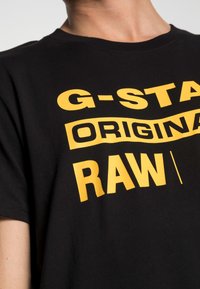 Camiseta de algodón negra con un atrevido texto amarillo que dice "G-STAD ORIGINAL RAW" en diferentes tamaños y estilos de fuente.