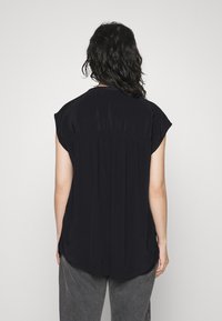 Top nero con maniche corte, leggermente oversize, caratterizzato da un dettaglio plissettato sul retro e realizzato in un tessuto morbido. L'orlo è arrotondato sui lati.