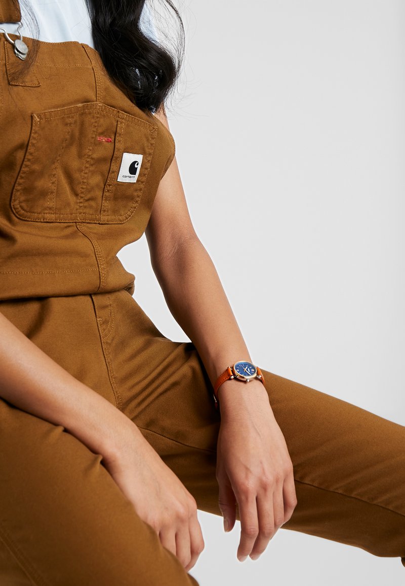 Brown overalls with a Carhartt logo pocket, worn with a light blue shirt and a watch featuring a blue face and an orange leather strap.