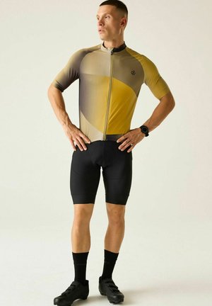 Atleet die een geel-grijs wielershirt draagt, zwarte gevoerde korte broek, zwarte sokken en wielrenschoenen, staand met de handen in de zij en kijkt naar rechts.