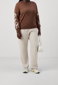 Felpa marrone con ricami floreali sulle maniche, abbinata a pantaloni beige chiaro su misura, borsa bianca e sneakers multicolori.