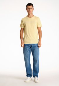 Gelbes Baumwoll-T-Shirt mit Rundhalsausschnitt und dem aufgedruckten Text "COPENHAGEN LINDHERGH", kombiniert mit blauen Jeans und weißen Sneakers.