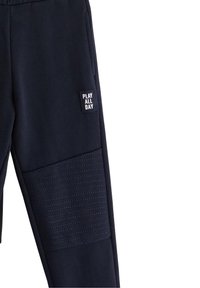 Οι μπλε ναυτικές φόρμες διαθέτουν υφασμάτινο πάνελ με οριζόντιες γραμμές, ελαστική μέση και τετράγωνο ένθετο με λευκό κείμενο που γράφει «ΠΑΙΞΕ ΟΛΗ ΜΕΡΑ».