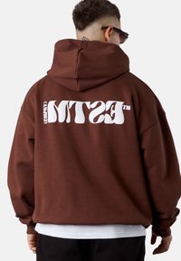 Hoodie surdimensionné marron avec un logo blanc au dos, texture douce, manches longues et grande capuche. Chemise blanche superposée visible en dessous.
