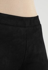 Pantaloni neri in finta suede con una trama liscia, dotati di una vita piatta e dettagli di cucitura sottili, mostrati sopra una maglietta beige chiaro.