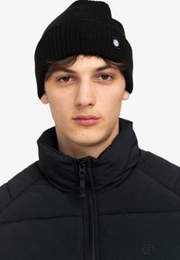 Beanie nero a coste con etichetta del logo, abbinato a un giubbino nero trapuntato con colletto alto e chiusura con zip.