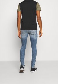 Camisa negra y oliva de manga corta, jeans azules de corte slim, y zapatillas negras con detalles en blanco. Presenta un corte clásico y denim desgastado.