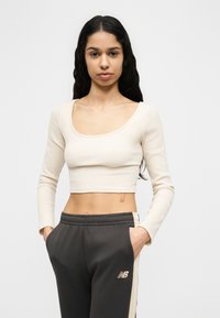 Νέα γυναίκα με μακριά σκούρα μαλλιά, φορώντας κρεμ χρωματιστό κοντό τοπ με μακριά μανίκια και μαύρο παντελόνι φόρμας New Balance με ρίγες στο πλάι, χέρια στις τσέπες.