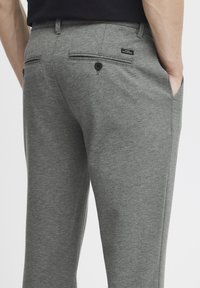 Pantalones elásticos grises con una textura suave, que cuentan con bolsillos traseros con botones y una etiqueta de logo, mostrados desde un ángulo lateral.