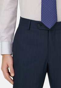 Mann trägt marineblaue Nadelstreifenhose, weißes Hemd mit dünnen vertikalen Streifen und blaue gemusterte Krawatte, Hand entspannt an der Seite.