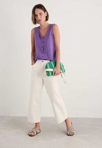 Purple sleeveless top with button details, paired with cream wide-leg pants. Carrying a green striped clutch bag; wearing gray sandals.