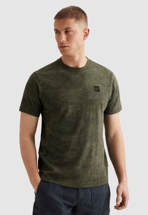 Muž v olivovo zelenej krátkorukávovej tričku s maskáčovým vzorom a námorníckych modrých cargo krátkych nohaviciach, pozerajúci sa doľava s rukami v vreckách.