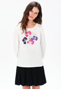 Witte langarmshirt met kleurrijke bloemenprint in roze, paars en marineblauw, voorzien van een ronde halslijn. Gekenmerkt door een zwarte plisseerrok.