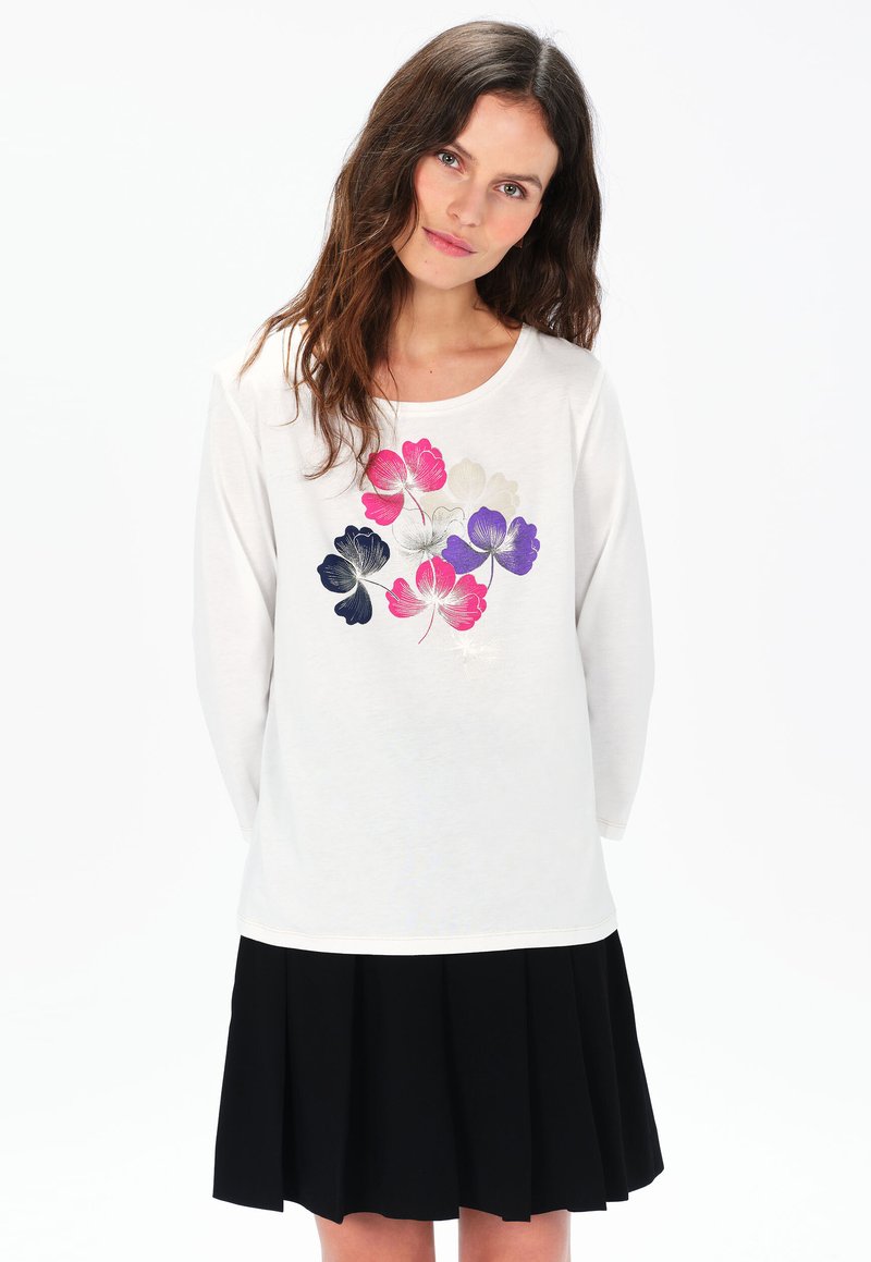 Weißes Langarmshirt mit buntem Blumenmuster in Rosa, Lila und Marineblau sowie rundem Ausschnitt. Kombiniert mit einem schwarzen Plisseerock.