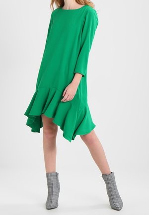 Kobieta ubrana w jaskrawą zieloną, asymetryczną sukienkę z falbanami, rękawami do łokcia i kratkowymi botkami, stojąca na jednolitym tle.