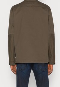 Brun bomuldjakke med lige snit, knaplukning ved ærmerne og sideslidser; båret over mørkeblå jeans. Enkel, minimalistisk design.