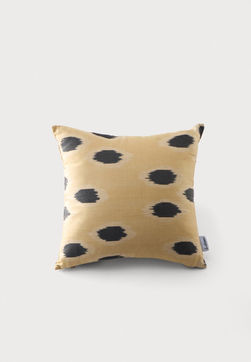 Coussin carré beige avec des taches noires irrégulières disposées en motif dispersé sur un fond uni.