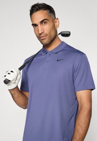 Fialová Nike polo košeľa s golierom, krátkymi rukávmi a logom na hrudi. Model drží golfovú palicu s bielou rukavicou.