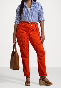 Cargo-byxor i klar orange tyg med dragkedjefickor, parat med en blå och vit randig skjorta med knappar och en brun handväska.