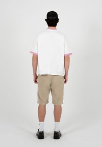 Mann trägt weißes T-Shirt mit pinkfarbenem Rand, beige Shorts, schwarze Kappe, weiße Socken und schwarze Schuhe, steht mit dem Rücken zum Betrachter vor weißem Hintergrund.