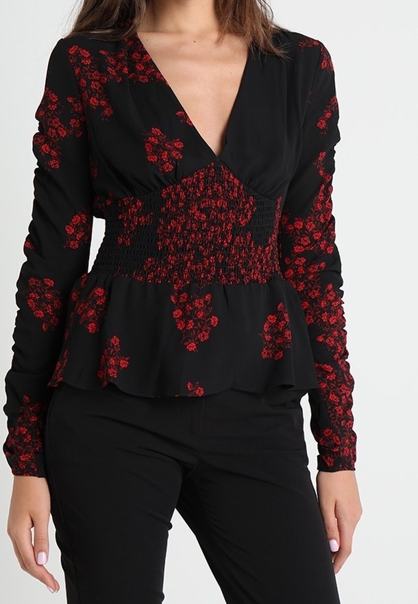 Femme portant un chemisier noir à manches longues avec des motifs floraux rouges, taille smockée et ourlet peplum, associé à un pantalon noir.