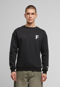 Must nääriga must kampsun, mille rinnal on valge "F" logo ja varrukal brändi aktsent. Pehme, tekstuuriga kanga.