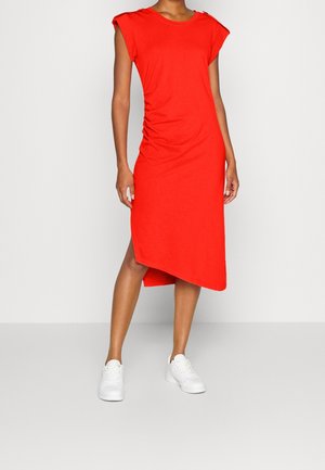 Kobieta w czerwonej, bezrękawnikowej sukience midi z asymetrycznym dołem i białych trampkach, stojąca na tle jednolitego tła.