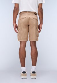Pantalones cortos de carga beige con una tela ligera, presentando bolsillos laterales y un corte relajado. Combinados con calcetas blancas y zapatillas neutras.