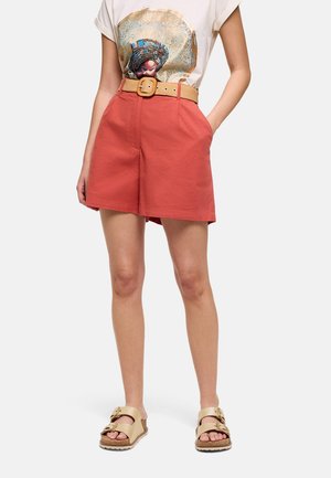 Mujer con pantalones cortos de color óxido con un cinturón beige tejido, una camiseta blanca con gráfico y sandalias beige con hebilla, de pie con las manos en los bolsillos.