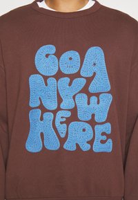 Felicino di colore marrone con lettering blu texturizzato a bolle che dice "GO ANYWHERE" in bella vista sul davanti.