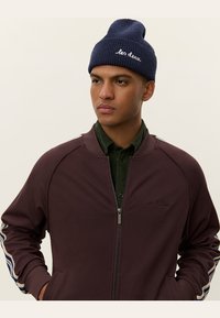 Jachetă zip-up în culoare mahon, cu manșete dintr-un material ribbed, având accente albe și albastre pe mâneci. Purtați peste o cămașă butonată de culoare verde închis. Căciulă bleumarin.