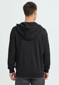 Sweat à capuche noir en tissu doux, avec une capuche doublée, des poignets et un ourlet côtelés. Le modèle présente une coupe décontractée avec une texture lisse.