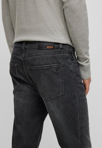 Person som bär svarta jeans i denim och en ljusgrå tröja, visar ryggen med synlig BOSS-logotyp på bälteslappen och en liten logotyp på fickan.