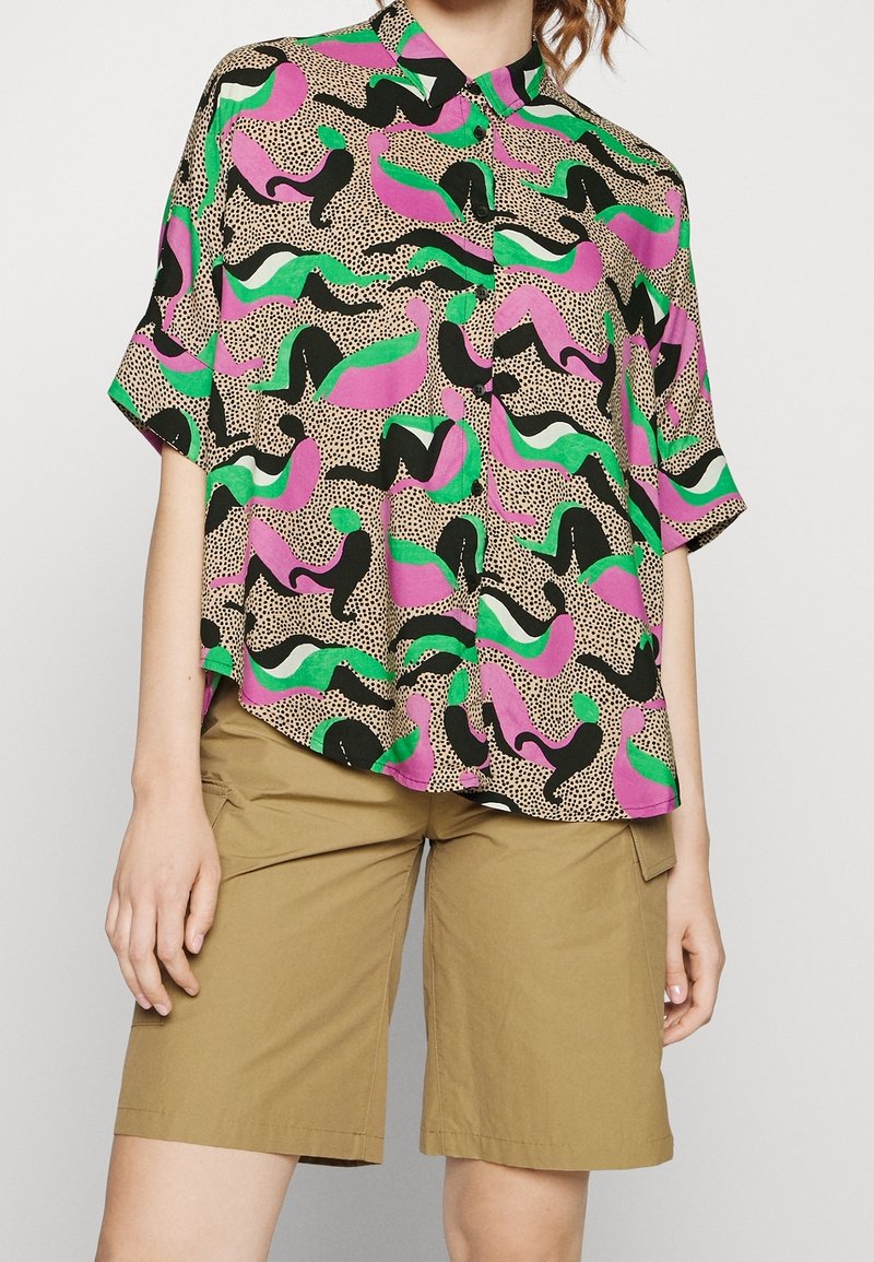 Camicia a manica corta dai colori vivaci con forme astratte in rosa, verde, nero e bianco su uno sfondo beige a pois.