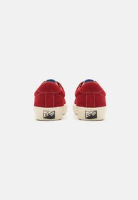 Red suede sneakers with a low profile, white rubber sole, blue inner lining, and logo on the heel. Rounded shape with stitched accents.