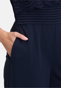 Main dans la poche d’un vêtement bleu marine avec un détail plissé à la taille et un tissu en dentelle sur le bord supérieur.