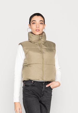 Chaleco de plumas en verde oliva, diseño acolchado, cuello alto, cierre frontal de cremallera. Lleva puesto sobre una camiseta blanca de manga larga con canalé y jeans oscuros.