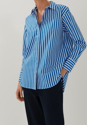 Person iført en blå og hvid lodret stribet skjorte med knapper og lange ærmer samt sorte bukser, den ene hånd i lommen.