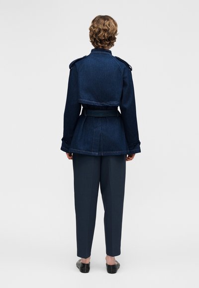 Άτομο με κοντά σγουρά μαλλιά που φοράει σκούρο μπλε σακάκι με ζώνη και επωμίδες και σκούρους μπλε παντελόνι, όρθιο με την πλάτη προς τα εμάς σε απλό φόντο.