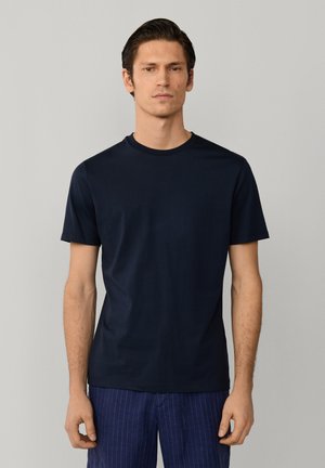 Homme portant un t-shirt à manches courtes bleu marine uni et un pantalon bleu à fines rayures, debout devant un fond gris clair.