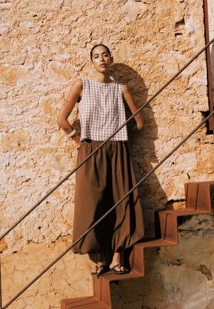 Kvinde i brune vide bukser og ternet ærmeløs top stående på rustne metaltrapper mod en struktureret stenvæg i sollys.
